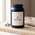 Anchor Whey Protein Isolate Vanilla navy tub on travertine stone with eucalyptus, natural window light