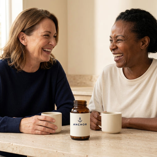 Two women in their 50s — one white, one Black — laughing over coffee at a kitchen table with an Anchor multivitamin amber bottle between them