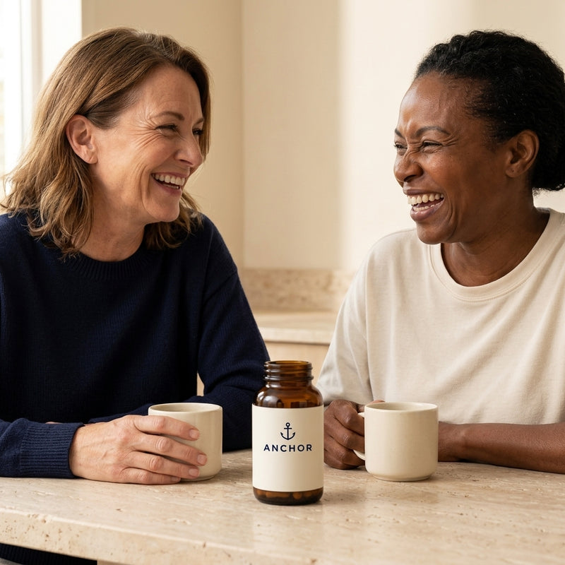 Two women in their 50s — one white, one Black — laughing over coffee at a kitchen table with an Anchor multivitamin amber bottle between them
